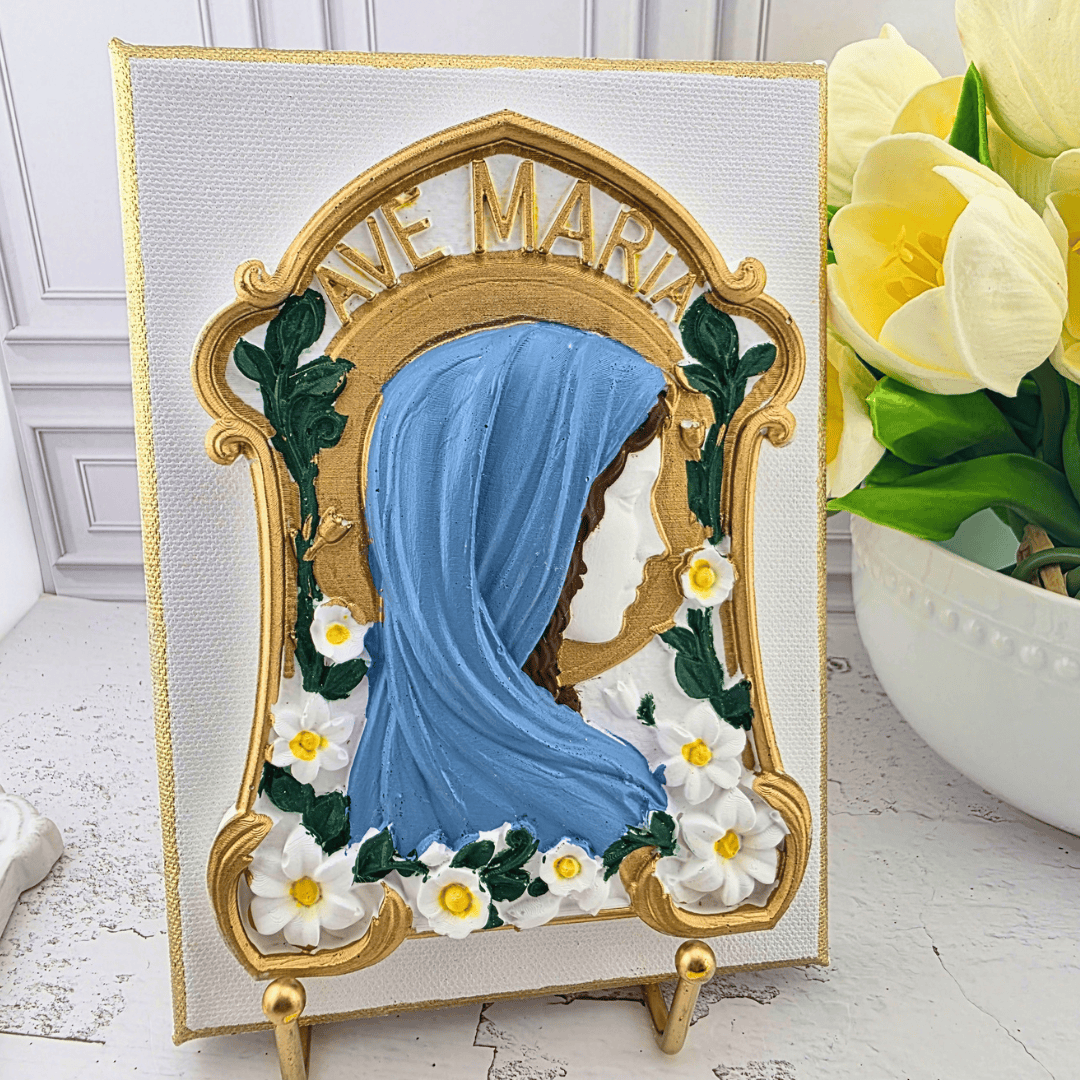 A decorative canvas art piece featuring a gold-framed portrait of the Virgin Mary with a blue veil, surrounded by white flowers, displayed on a small gold stand, Sister Dulce Gift Shop, Shepherds Staff, Religious Store, Cypress Springs Mercedarian Prayer Center, Catholic,