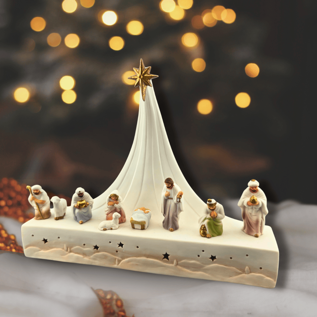 Sister Dulce Gift Shop, Shepherds Staff, Religious Store, Cypress Springs Mercedarian Prayer Center, Catholic, A ceramic Christmas nativity scene figurine set on a rectangular base with a tall white tent, gold star, and small detailed characters in soft pastel colors, photographed against a dark blurred background with warm bokeh lights.