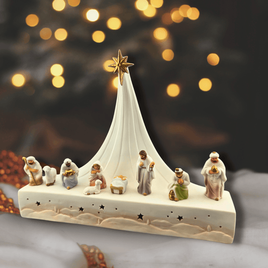 Sister Dulce Gift Shop, Shepherds Staff, Religious Store, Cypress Springs Mercedarian Prayer Center, Catholic, A ceramic Christmas nativity scene figurine set on a rectangular base with a tall white tent, gold star, and small detailed characters in soft pastel colors, photographed against a dark blurred background with warm bokeh lights.