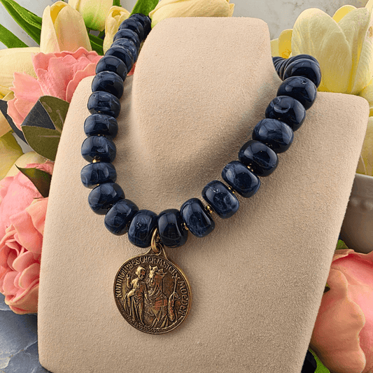 Sister Dulce Gift Shop, Shepherds Staff, Religious Store, Cypress Springs Mercedarian Prayer Center, Catholic, A beaded necklace made of dark navy blue round beads with a bronze-toned circular pendant, displayed on a beige jewelry bust surrounded by pink and yellow flowers.