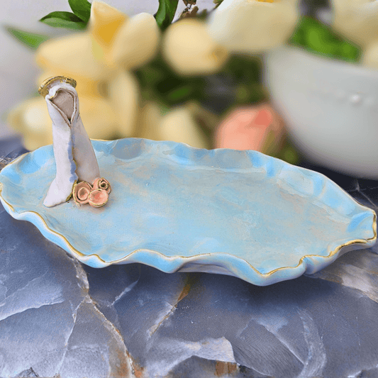 Sister Dulce Gift Shop, Shepherds Staff, Religious Store, Cypress Springs Mercedarian Prayer Center, Catholic,A light blue ceramic dish with a wavy leaf-like edge and gold trim, holding a miniature white dress and pink shoes. The dish is styled on a gray stone surface with blurred cream-colored flowers in the background.