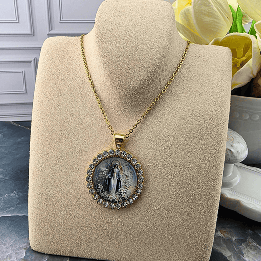 A gold chain necklace with a round pendant featuring a religious image of the Virgin Mary, displayed on a beige velvet jewelry bust. The pendant is bordered with small clear rhinestones, Sister Dulce Gift Shop, Shepherds Staff, Religious Store, Cypress Springs Mercedarian Prayer Center, Catholic, 