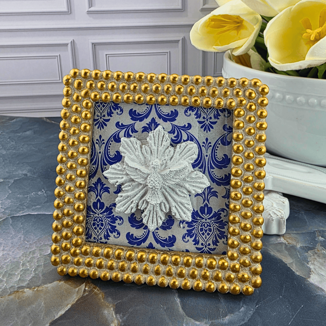 A square decorative picture frame with a gold beaded border and a blue-and-white damask patterned insert, featuring a white sculpted flower in the center. Sister Dulce Gift Shop, Shepherds Staff, Religious Store, Cypress Springs Mercedarian Prayer Center, Catholic, 