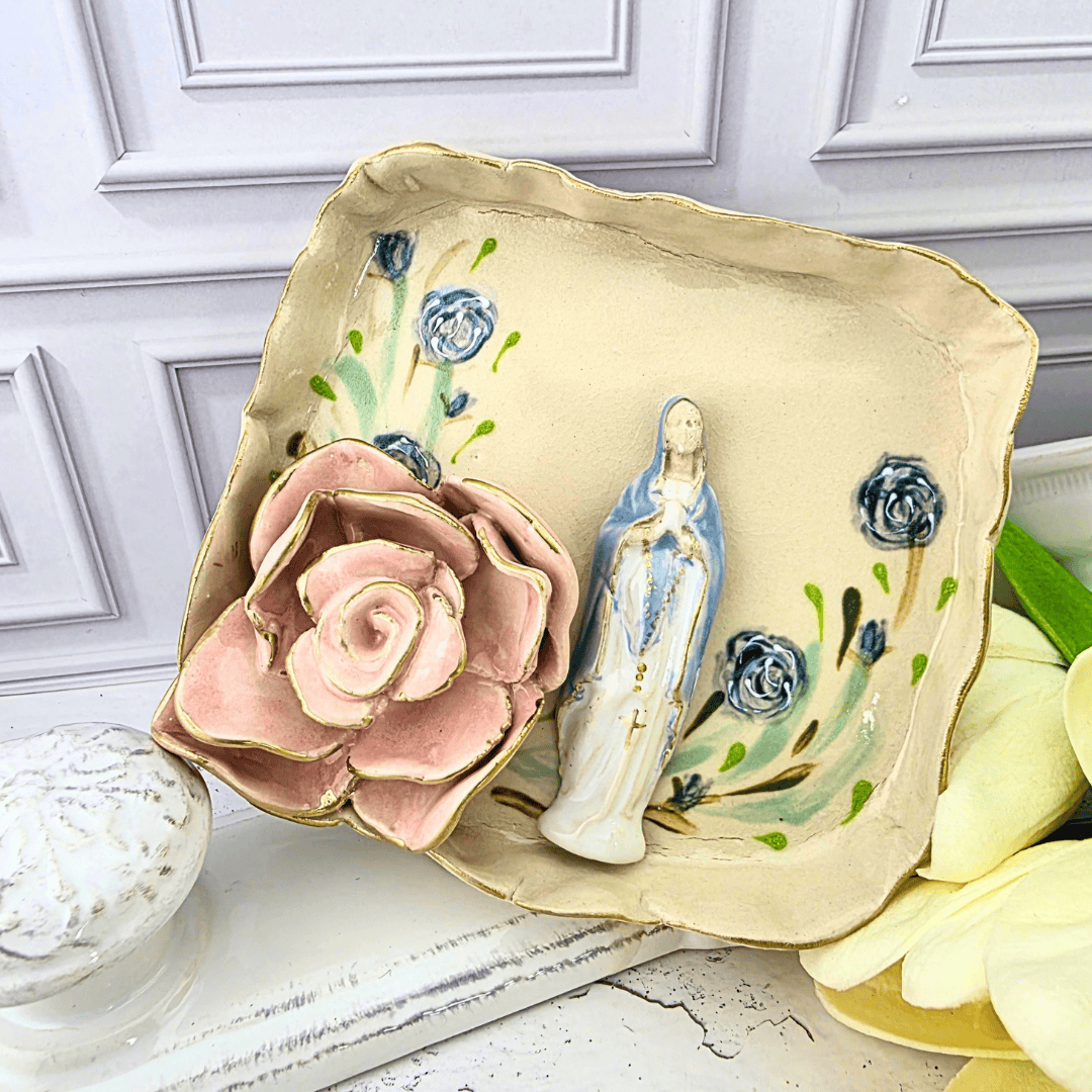 A decorative ceramic dish with a scalloped cream border and gold trim, featuring painted blue flowers, green leaves, a pink rose, and a small blue-and-white figurine, our lady of the rosary, sacred treasures, Sister Dulce Gift Shop, Shepherds Staff, Religious Store, Cypress Springs Mercedarian Prayer Center, Catholic, new item, popular item, trinket dish, handmade, 