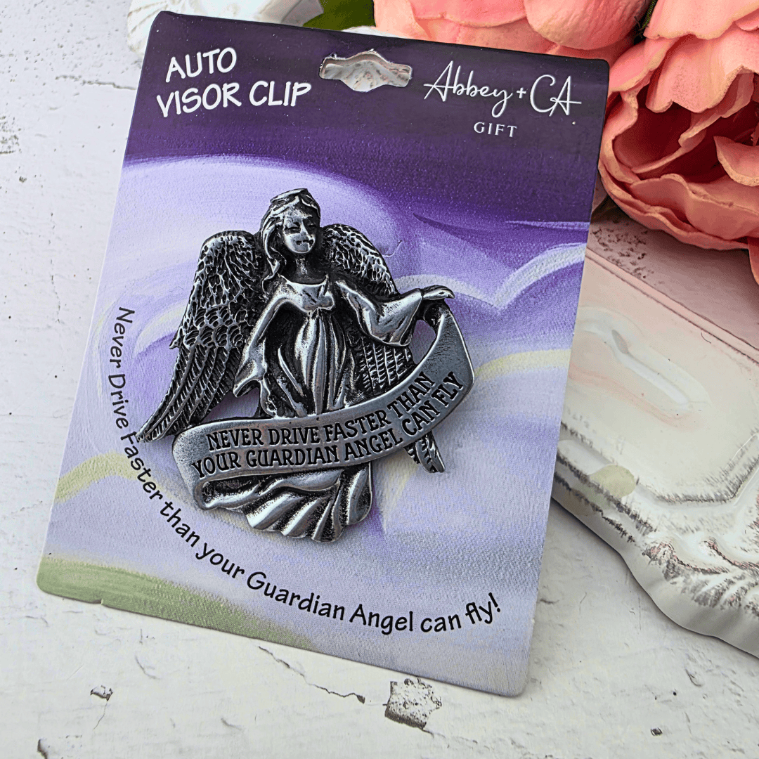 A silver-toned guardian angel visor clip mounted on a branded gift card, with the clip featuring an angel holding a banner that says, 'NEVER DRIVE FASTER THAN YOUR GUARDIAN ANGEL CAN FLY!', Sister Dulce Gift Shop, Shepherds Staff, Religious Store, Cypress Springs Mercedarian Prayer Center, Catholic, new item, young adult, new driver